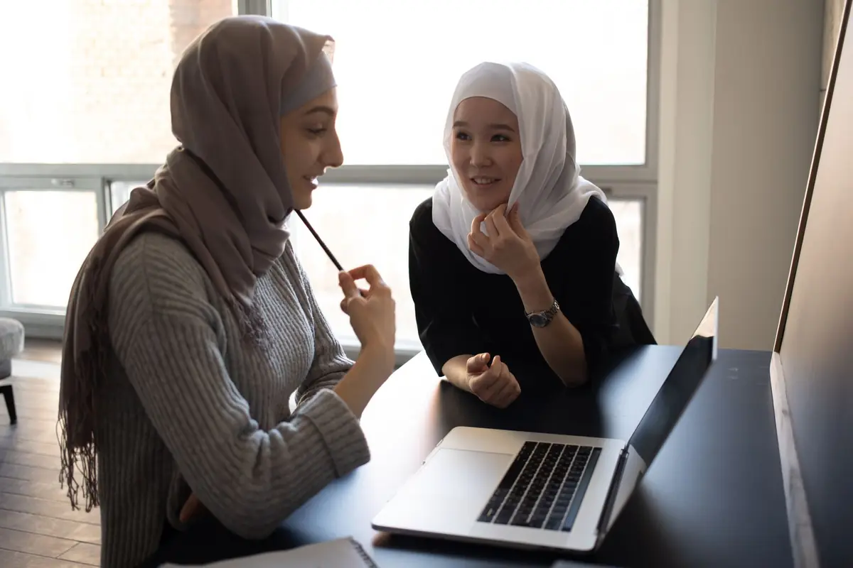 Dua staf administrasi berkerudung berdiskusi di depan laptop