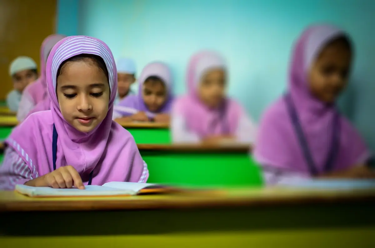 Siswi berkerudung ungu membaca di kelas, teman-temannya tampak kabur di belakang